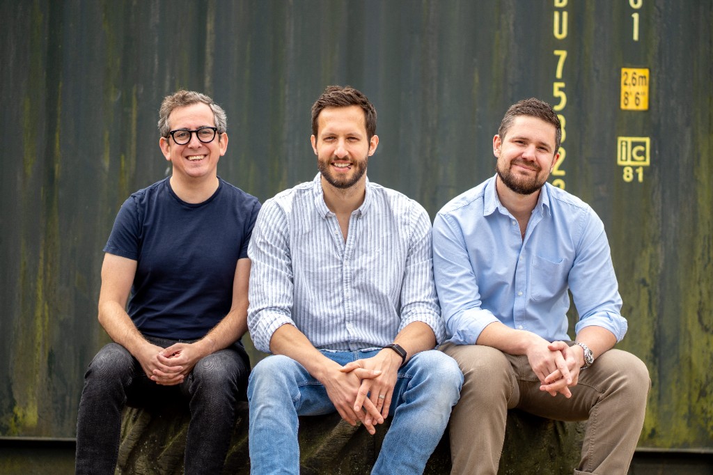 EngineLab founders, from left to right Matthew Herman, Daniel Goller and Sam Reid, sitting side by side in front of a weathered green shipping container.